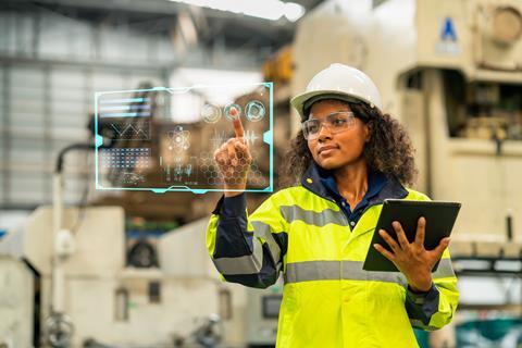 An engineer wearing safety gear in an industrial setting interacts with a futuristic screen
