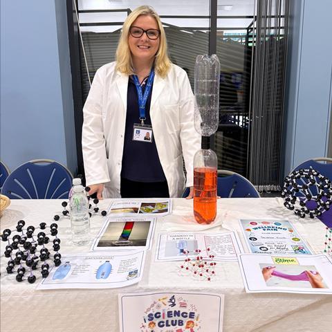 Lynsey Findlay wearing a lab coat manning the science club table at a school fair. There are posters, molecular models and science experiments on the table.