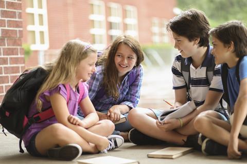 A picture showing two girls and two boys sitting on the ground talking, all wearing backpacks, against the backdrop of a building