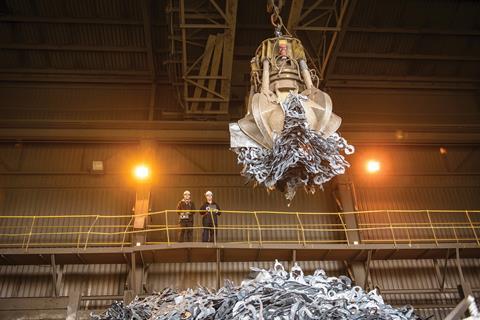 Two men control a mechanical claw to grab scrap metal at a recycling plant