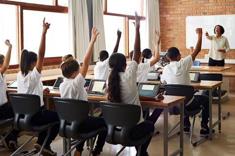 A photo of a teacher in front of a class of young students with their arms raised and tablet computers
