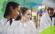 A female teacher and two students wearing white lab coats smile while observing apparatus set up for an experiment