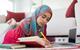 A female student wearing a hijab studies at home, using a notebook, pen and laptop