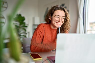 A young woman smiles while working at her desk in a home environment, looking towards her laptop