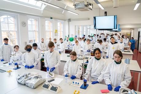Groups of students in white lab coats stand at work benches in a large school laboratory, looking towards the front and at their apparatus