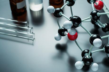 Close-up of a molecular model with black, white, and red atoms connected by rods beside glass test tubes and laboratory bottles on a lab bench.